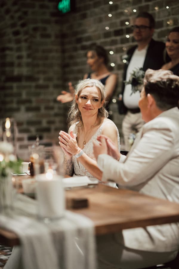 Bride Mandy and bride Selina sit at a table clapping during the reception at Sandstone Point Hotel — The Cellar, with guests visible in the background.