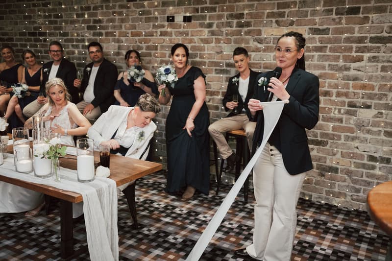 One bride stands and reads from a long scroll into a microphone at the Sandstone Point Hotel — The Cellar, while the other bride and guests seated nearby listen attentively.