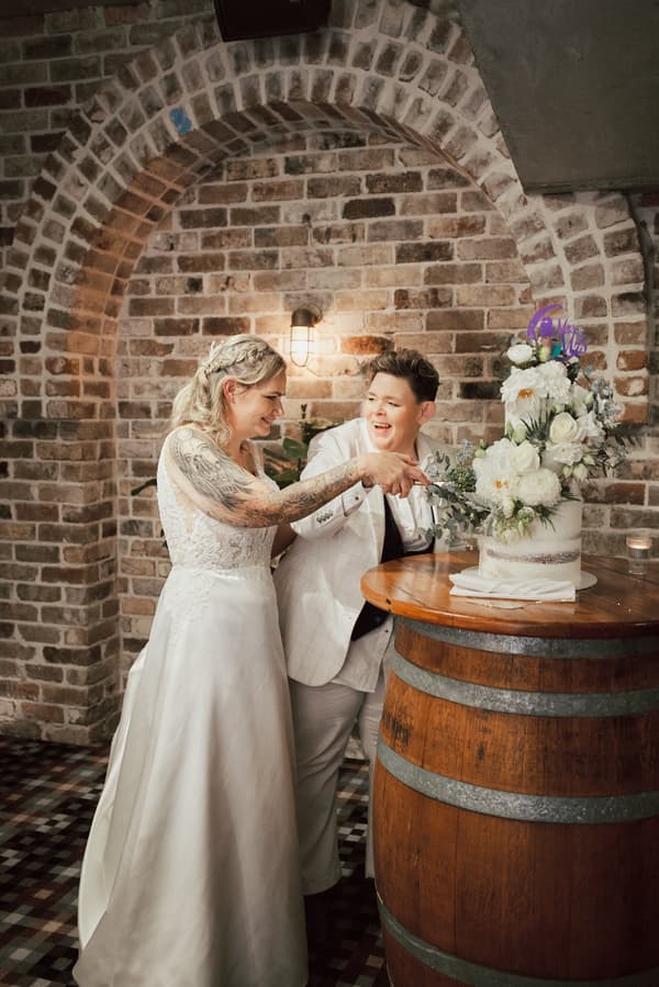 Brides Mandy and Selina cut their wedding cake together at Sandstone Point Hotel — The Cellar, standing by a wooden barrel table against a brick arch wall.