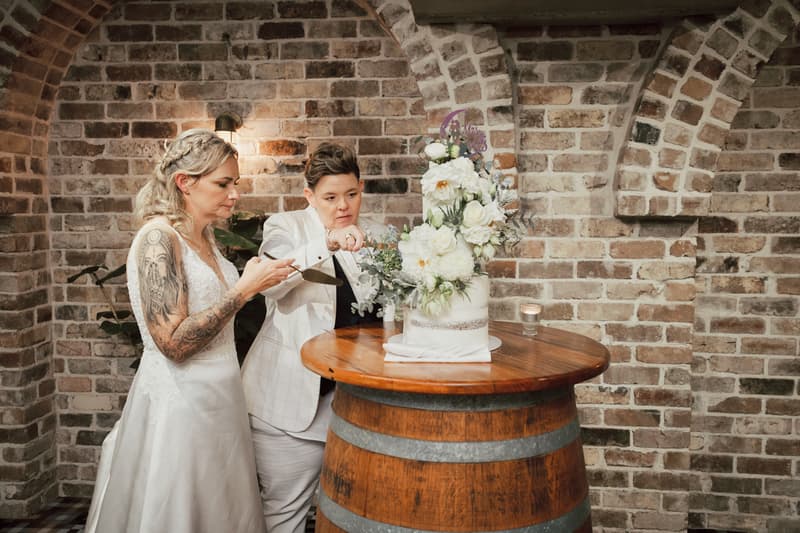 Brides Mandy and Selina cut their wedding cake on a wooden barrel table at Sandstone Point Hotel — The Cellar, surrounded by exposed brick arches.