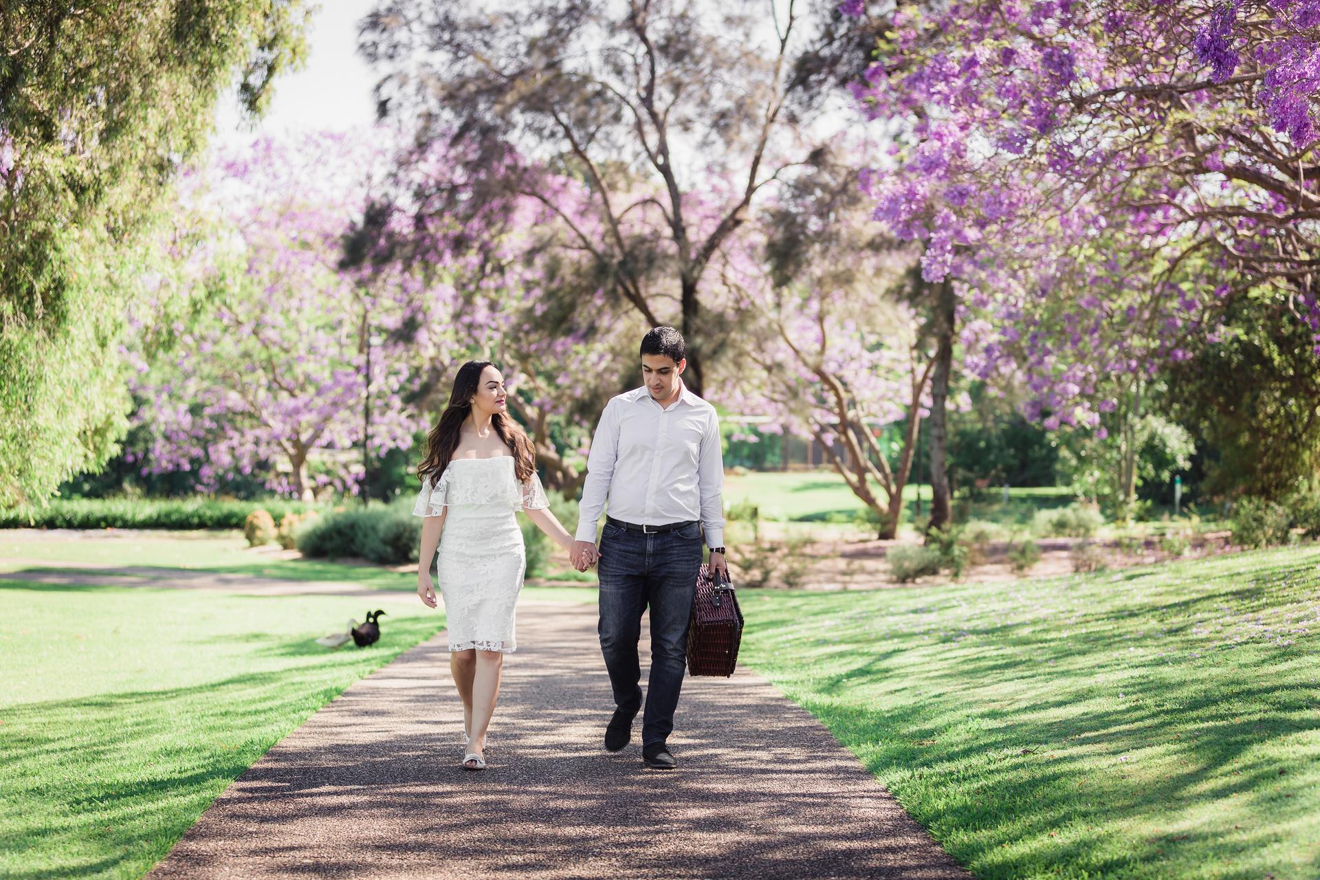 The couple walks hand in hand along a path at UQ Lakes, surrounded by lush greenery and purple blossoms.