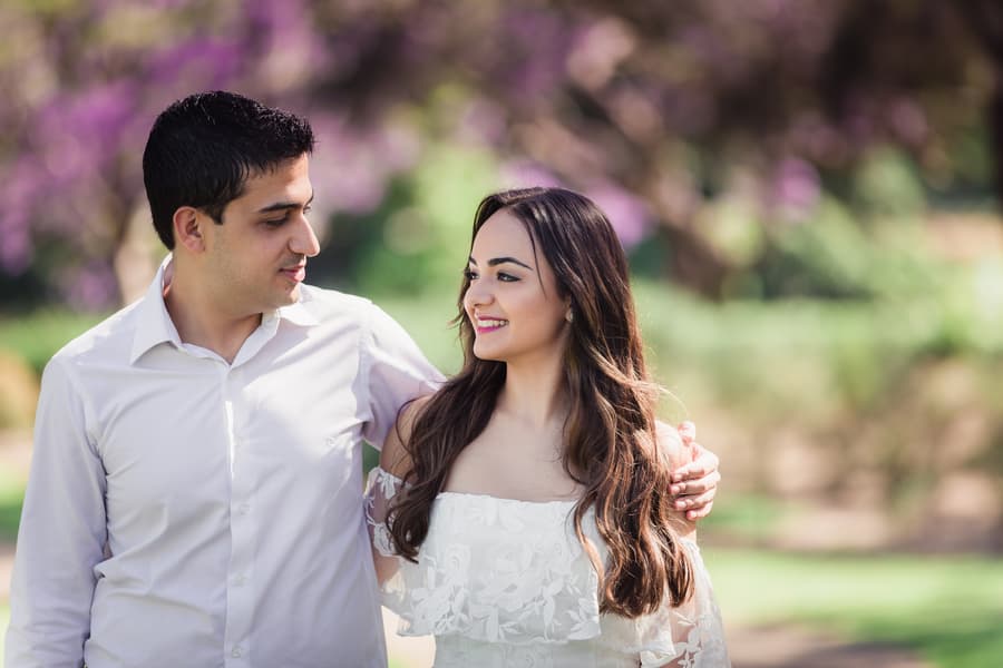 The couple gazes at each other lovingly, standing close with a backdrop of purple flowers at UQ Lakes.
