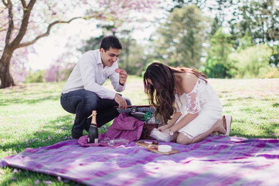 The couple enjoys a picnic on a purple blanket at UQ Lakes, with a basket and champagne nearby.