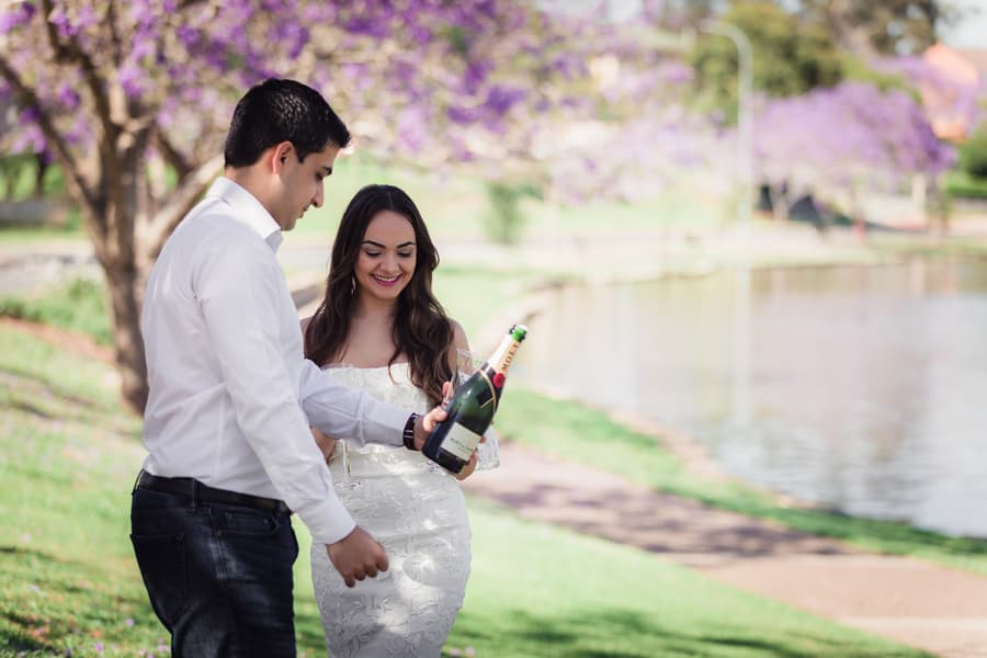 The couple stands by the lake at UQ Lakes, preparing to open a bottle of champagne together.