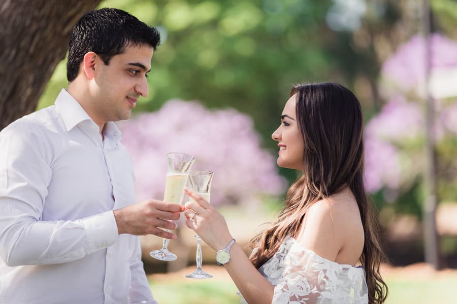 The couple clinks glasses of champagne, smiling at each other under the trees at UQ Lakes.