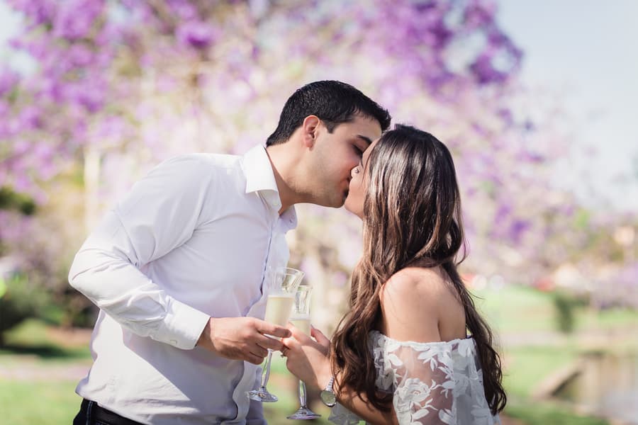 The couple shares a kiss while holding champagne glasses, with purple blossoms in the background at UQ Lakes.