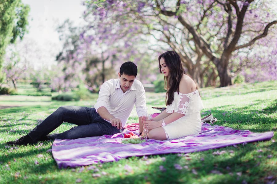 The couple relaxes on a picnic blanket, enjoying snacks together under the shade of trees at UQ Lakes.