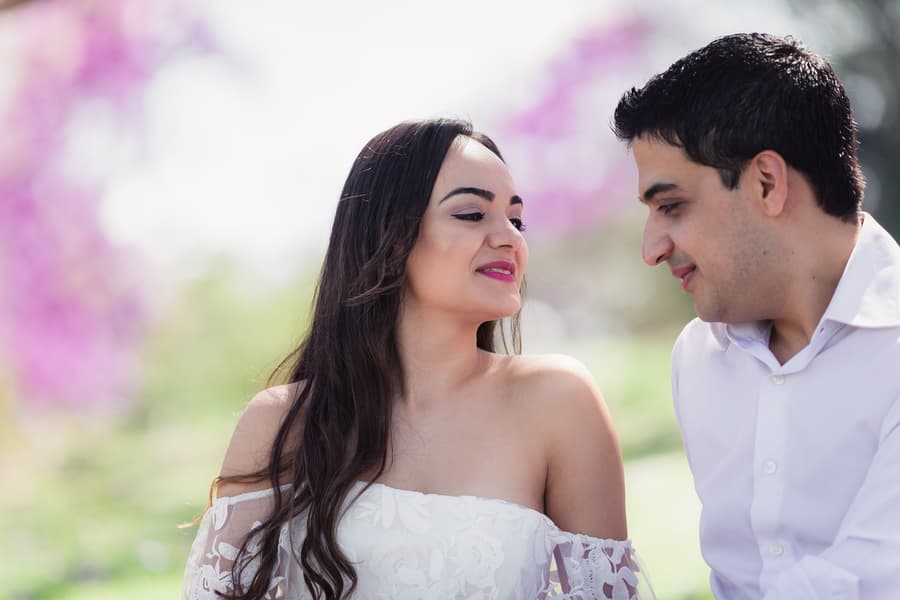 The couple sits close, exchanging affectionate looks, surrounded by greenery and purple flowers at UQ Lakes.