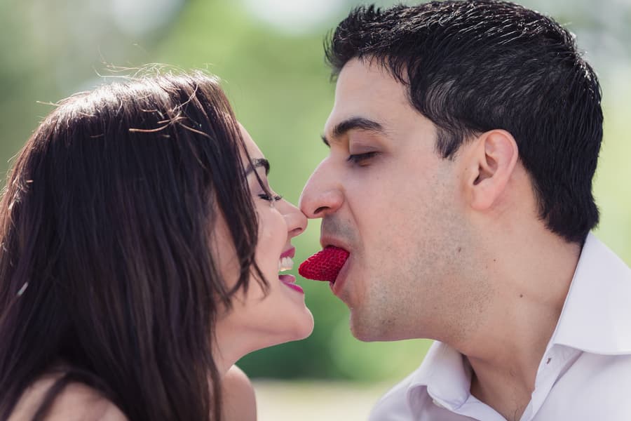 The couple playfully shares a strawberry, leaning towards each other with joyful expressions at UQ Lakes.