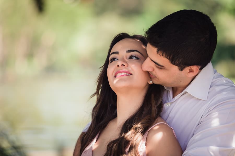 The couple shares an intimate moment, with one leaning in to kiss the other's cheek, surrounded by greenery.