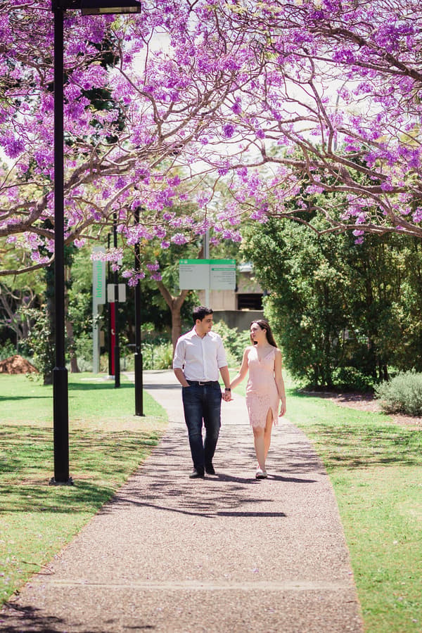 The couple walks hand in hand under blooming purple trees, enjoying a sunny day at UQ Lakes.