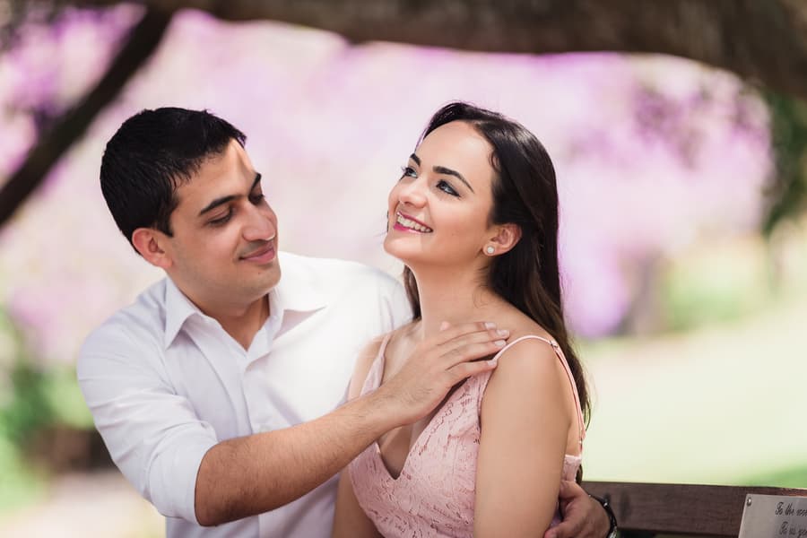 The couple sits on a bench, smiling at each other, with purple blossoms in the background at UQ Lakes.