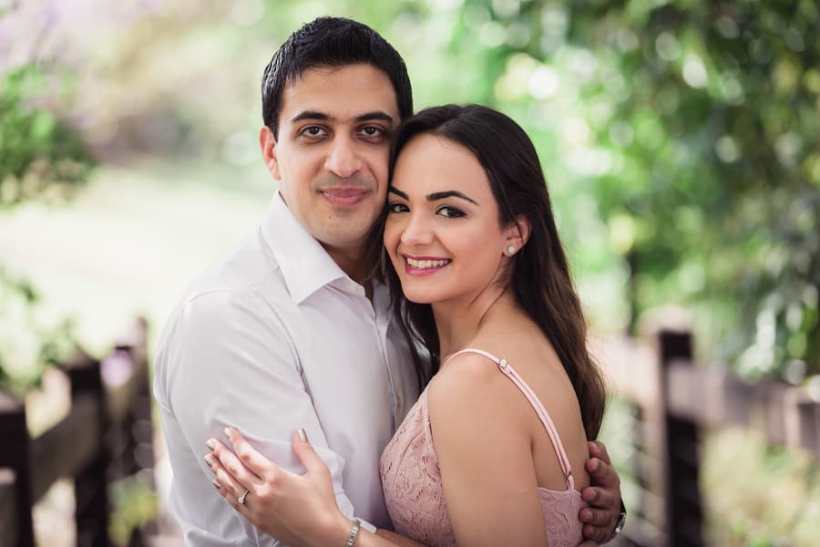 The couple stands close, smiling warmly at the camera, with a lush green backdrop at UQ Lakes.