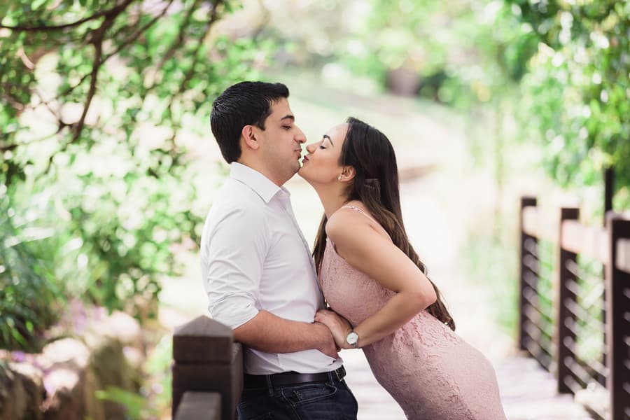 The couple leans in for a kiss on a wooden bridge, surrounded by vibrant greenery at UQ Lakes.