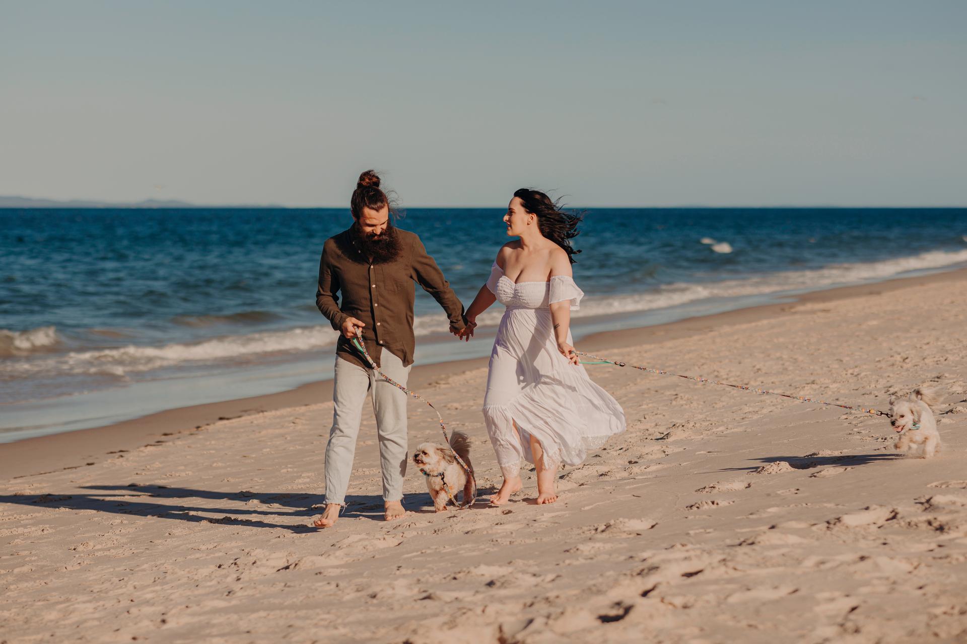 The couple walks barefoot on Woorim Beach, holding hands with two small dogs on leashes. The sun casts long shadows on the sand.