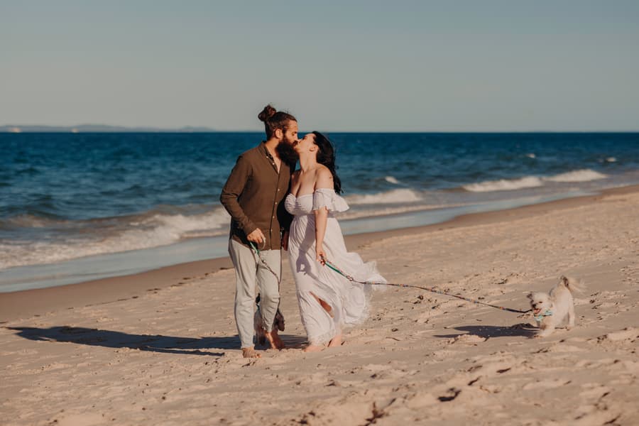 The couple shares a kiss on Woorim Beach, with their dogs playfully pulling on their leashes. The ocean waves gently roll in the background.