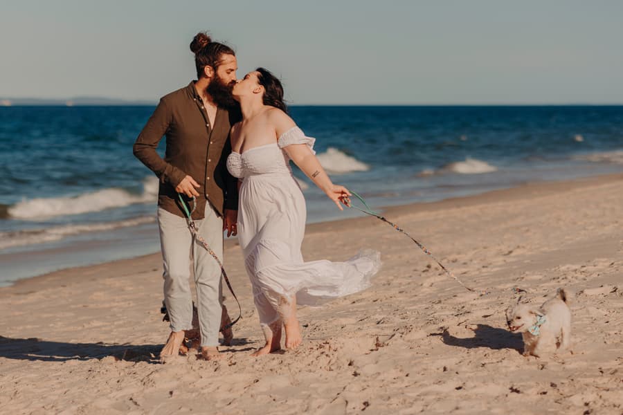 The couple kisses on the beach, holding their dogs' leashes. The sand and sea create a serene backdrop.
