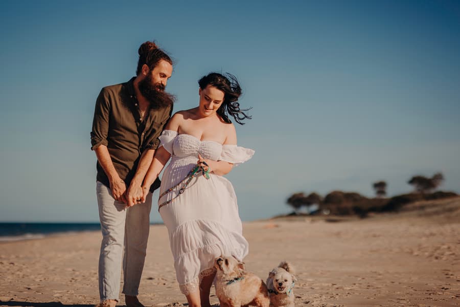 The couple walks hand in hand on the beach, smiling at each other with their dogs at their feet. The sky is clear and blue.