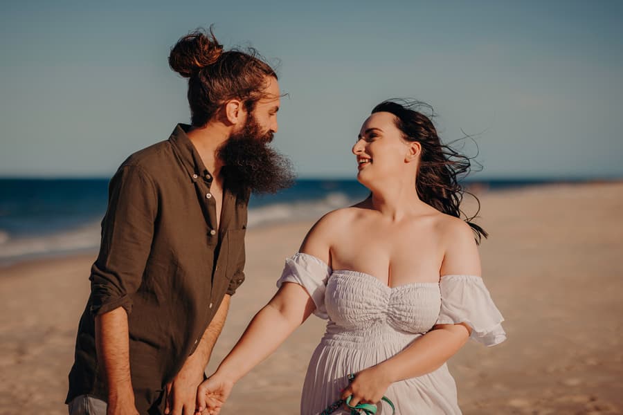 The couple stands close, smiling warmly at each other on the beach. Their dogs sit contentedly by their feet.