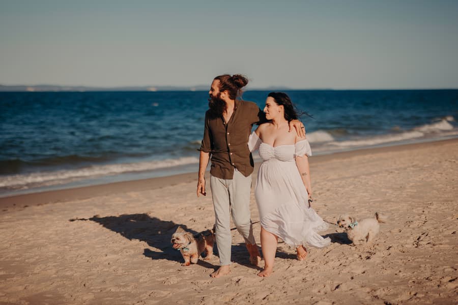 The couple gazes into each other's eyes, holding hands on the sandy beach. The sunlight highlights their happy expressions.