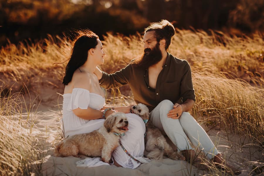 The couple sits on the sand dunes at Banksia Beach, surrounded by tall grass. They smile at each other with their dogs nearby.