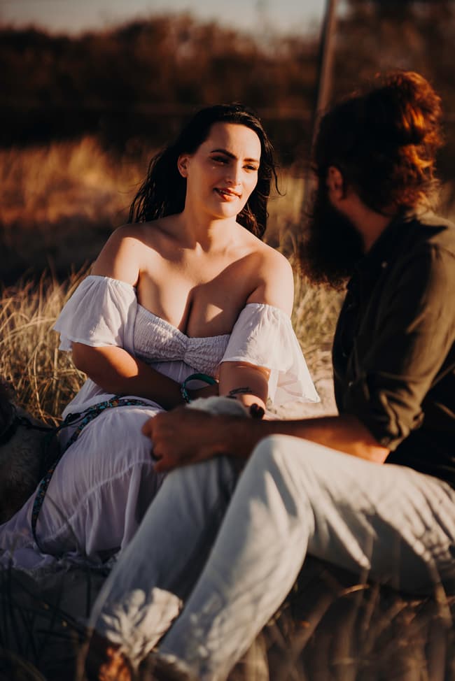 The couple sits close together on the sand, sharing a tender moment with their dogs. The grass sways gently in the breeze.
