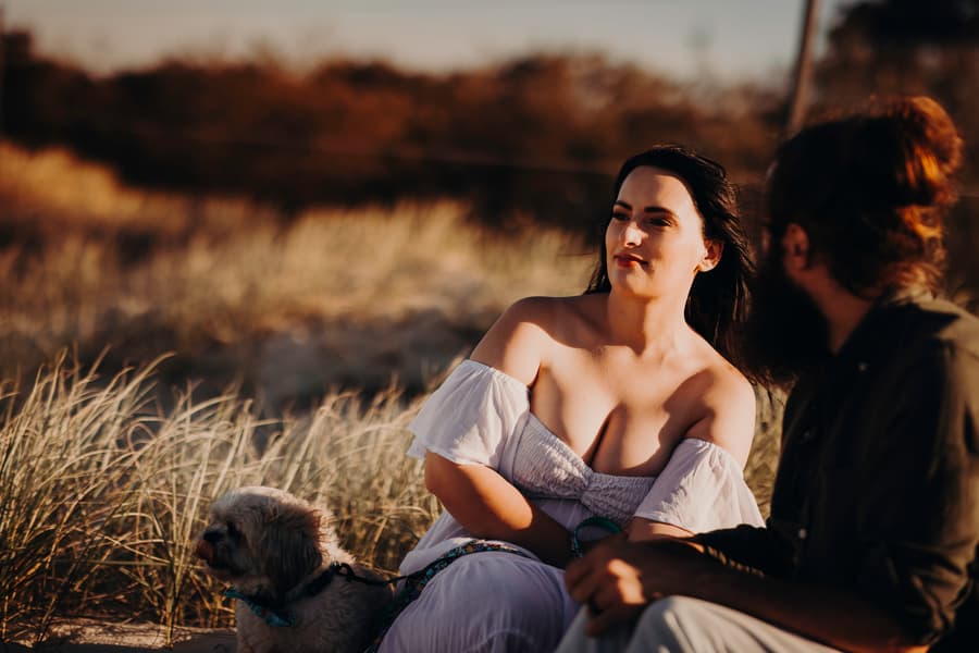 The couple sits facing each other on the sand, engaged in conversation. The light is soft and warm, creating an intimate atmosphere.
