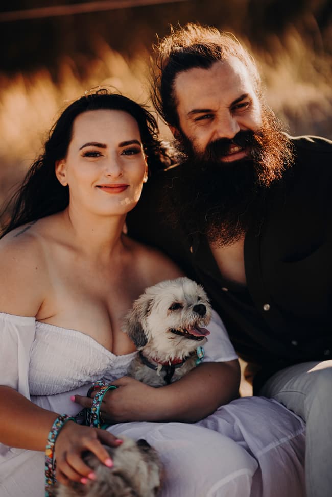 The couple sits together on the sand, with one of their dogs resting beside them. The setting sun casts a golden glow.