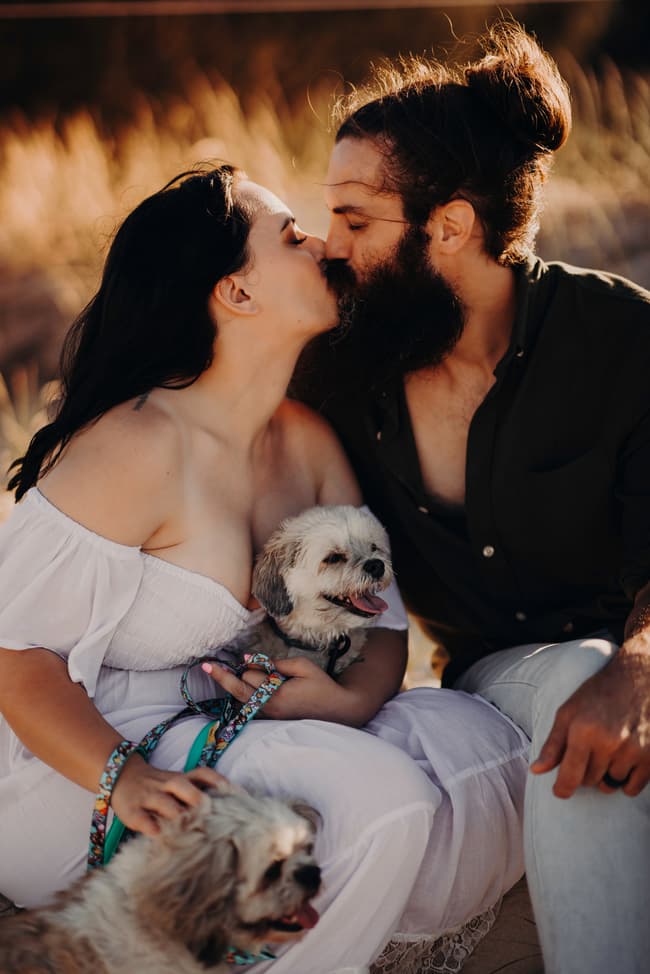 The couple sits closely, smiling with their dogs in their laps. The grass and sand create a natural setting.