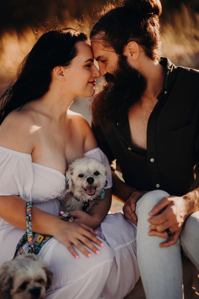 The couple sits closely on the sand at Woorim Beach, gently touching foreheads, with two small dogs in their laps. The sunlight creates a warm, intimate atmosphere.