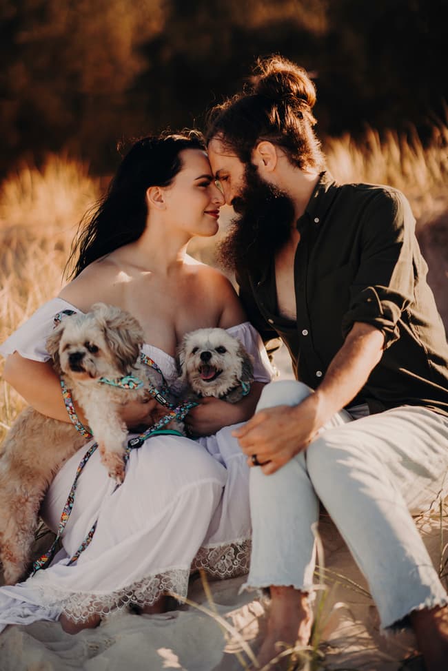 The couple sits on the sand at Woorim Beach, holding two small dogs. They touch foreheads, surrounded by tall grass, in a serene setting.