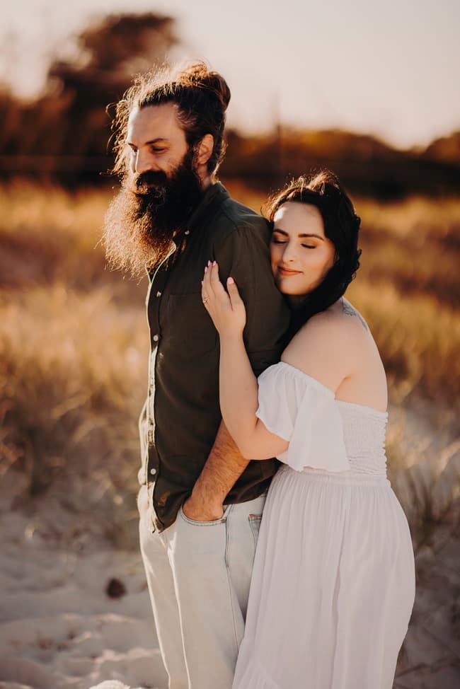 The couple stands on the sand at Woorim Beach, with one embracing the other from behind. The warm sunlight highlights their relaxed expressions.