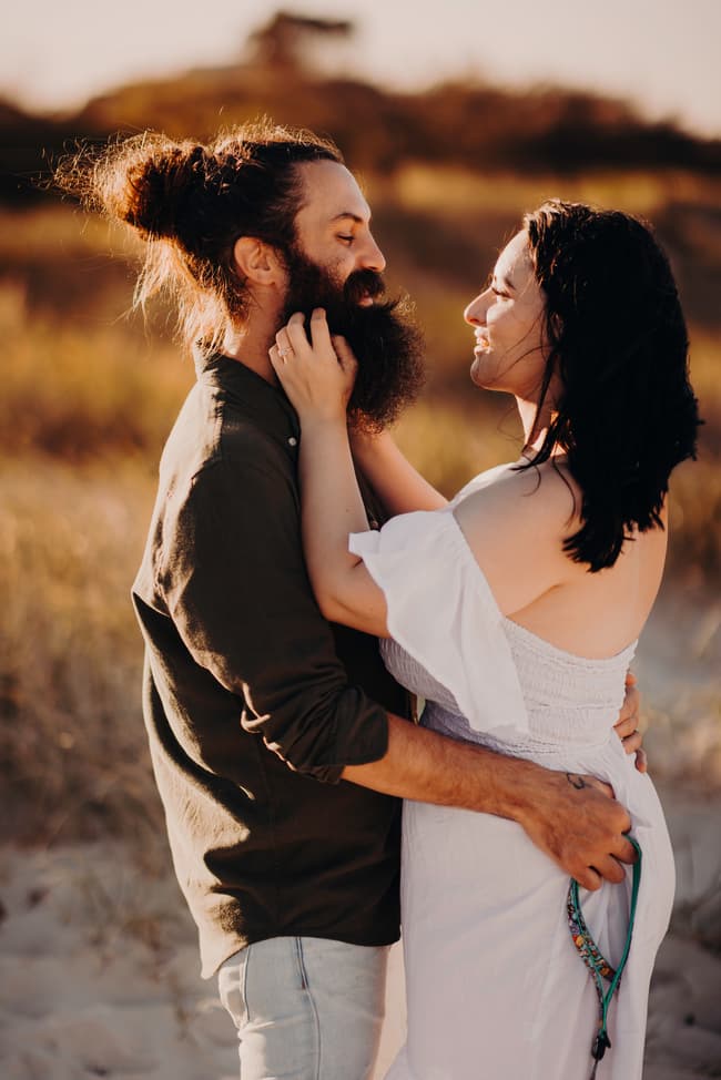 The couple stands facing each other on the sand at Woorim Beach, smiling warmly. The sunlight casts a gentle glow on their faces.