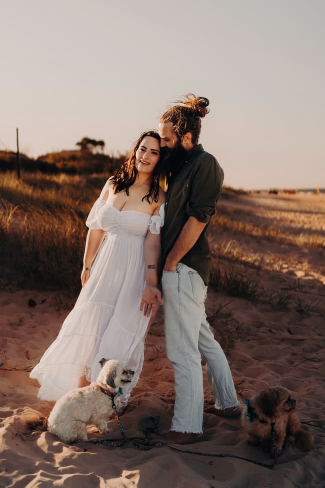 The couple stands on the sand at Woorim Beach, with one leaning on the other. Two small dogs sit at their feet, and the setting sun casts a warm glow.
