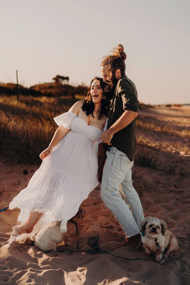 The couple stands playfully on the sand at Woorim Beach, laughing together. Two small dogs are by their side, adding to the joyful scene.