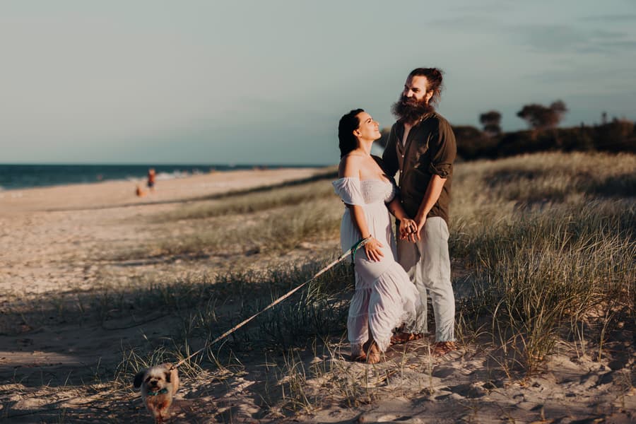 The couple stands on the sandy path at Woorim Beach, holding hands and smiling. The ocean is visible in the background, adding to the serene mood.