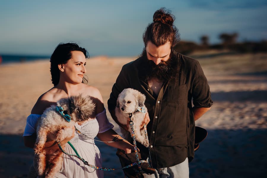 The couple stands on the sand at Woorim Beach, holding two small dogs. They look at each other with affection, under the soft beach light.
