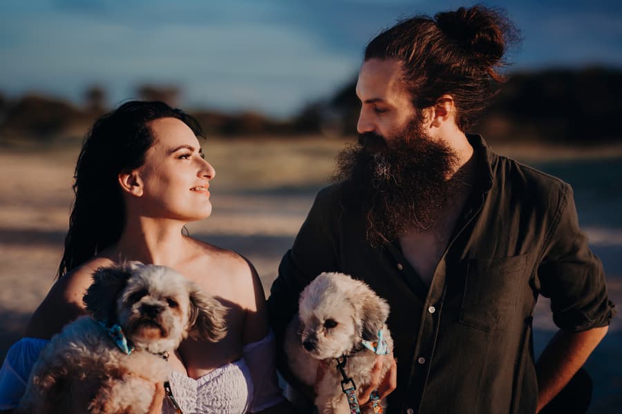 The couple walks along the shoreline at Woorim Beach, holding two small dogs. They look at each other with smiles, enjoying the beach setting.