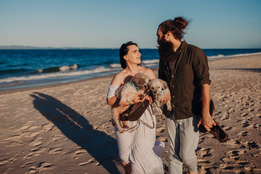 The couple walks hand in hand along the beach at Woorim Beach, carrying shoes. The ocean waves gently lap at the shore, creating a peaceful scene.