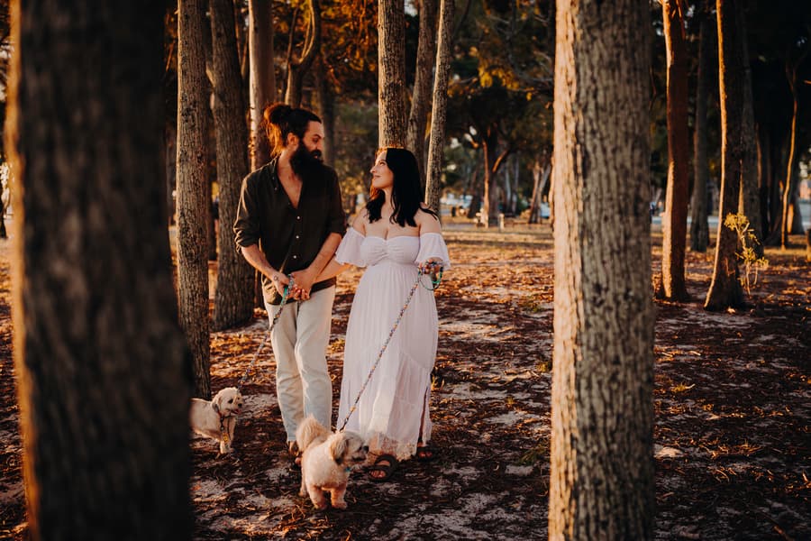 The couple walks through a sunlit forest at Woorim Beach, holding hands and leading two small dogs.