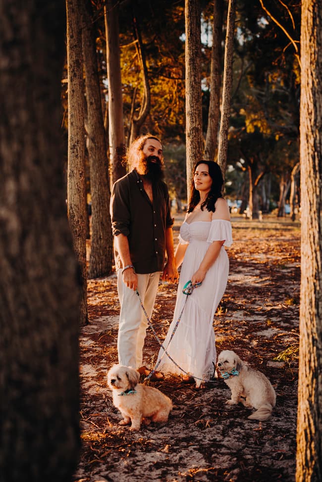 The couple stands closely in a wooded area at Woorim Beach, with two dogs on leashes, looking content.