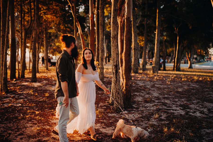 The couple walks through a forest at Woorim Beach, with the woman looking up and smiling, holding a dog leash.