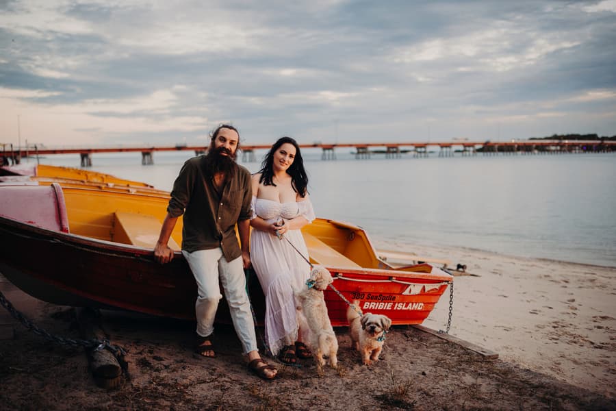 The couple poses with their dogs by a boat on the shore at Bribie Island, looking relaxed.