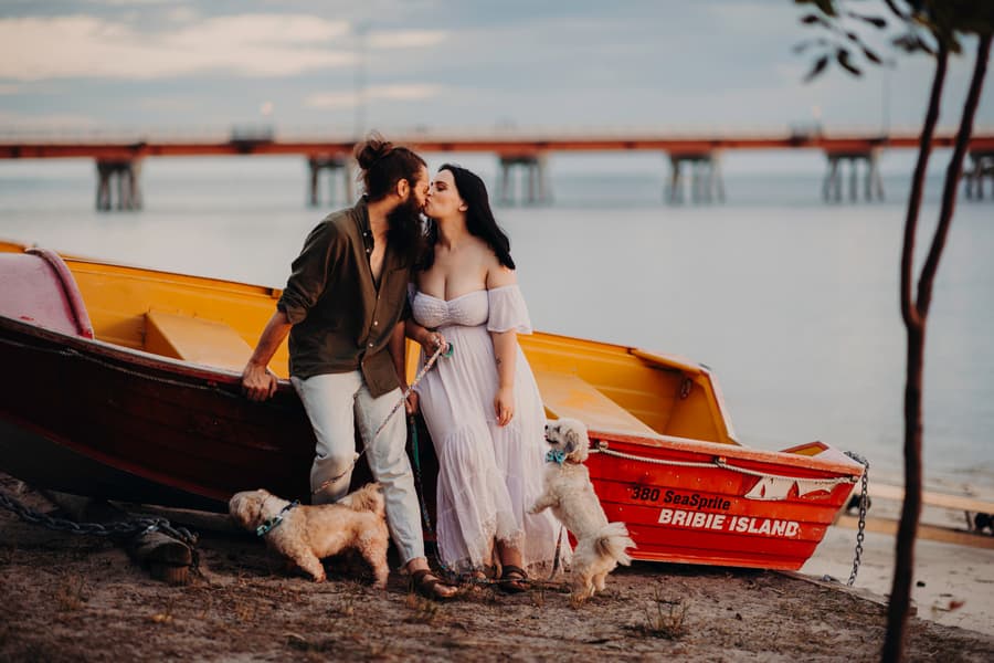 The couple shares a kiss by a boat on the shore at Bribie Island, with their dogs nearby.