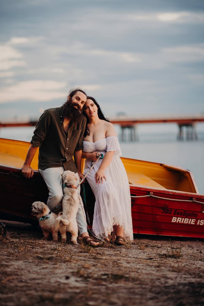 The couple leans against a boat on the shore at Bribie Island, with their dogs at their feet.