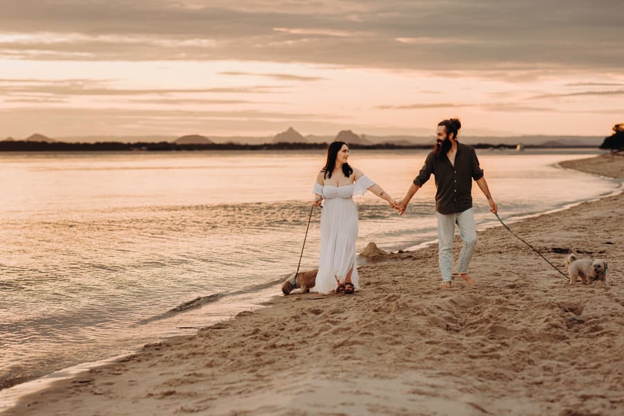 The couple walks hand in hand along the beach at sunset, with their dogs leading the way.