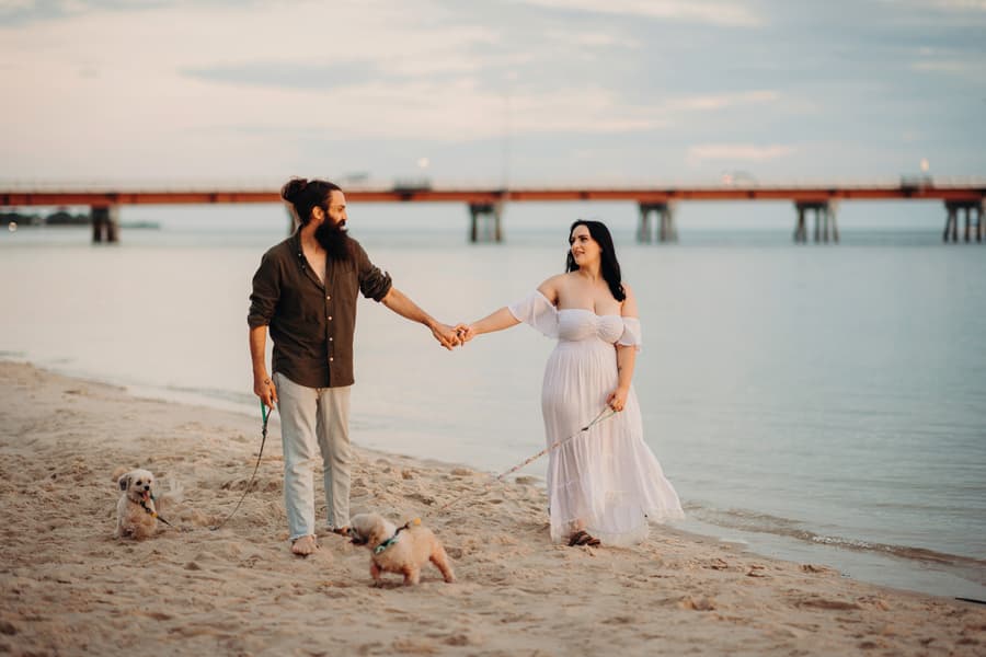 The couple holding hands and walking along the sandy shore of Woorim Beach, each holding a small dog on a leash. The sky is overcast.