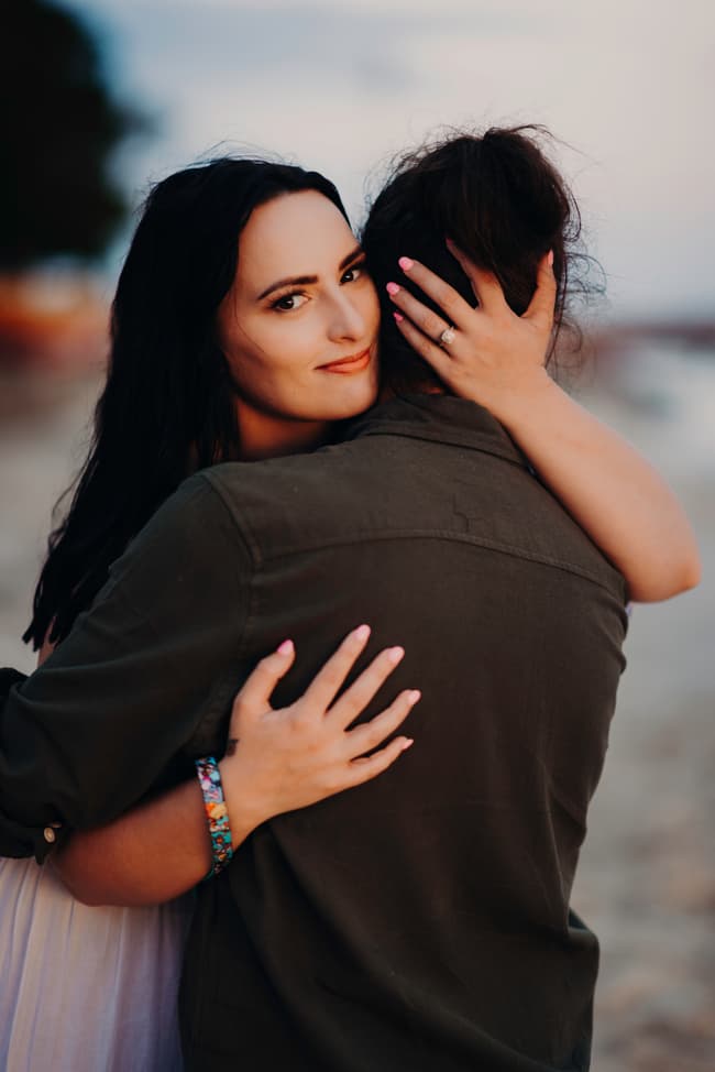 One person embracing the other from behind on the beach, showing an engagement ring. The mood is intimate and joyful.