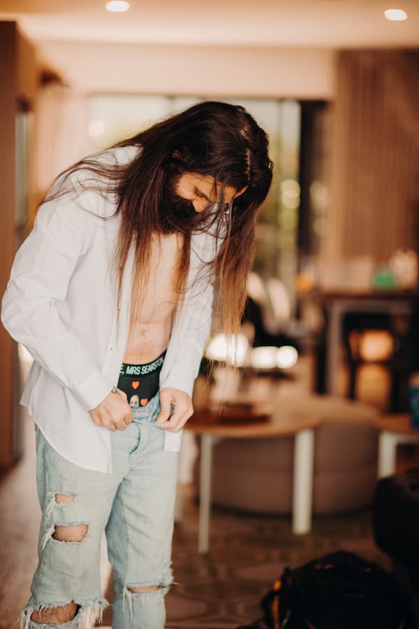 The groom David is buttoning his jeans while wearing an unbuttoned white shirt and visible black underwear with text at Sandstone Point Hotel.