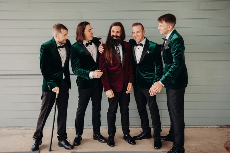 David with four groomsmen wearing green velvet jackets and black bow ties, standing against a light gray wooden wall at Sandstone Point Hotel.